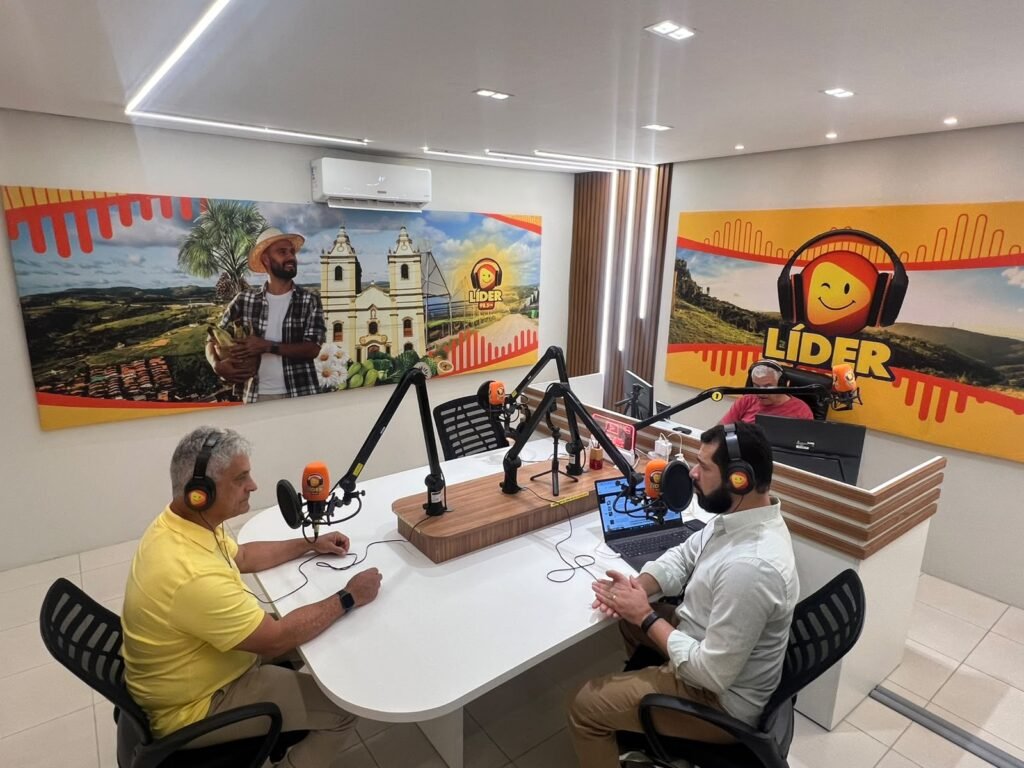 Um homem de camisa polo amarela conversa com um homem de camisa de botão clara em um estúdio de rádio. O homem de amarelo gesticula e o homem de claro escuta. Eles estão em uma mesa branca com microfones e fones de ouvido.
