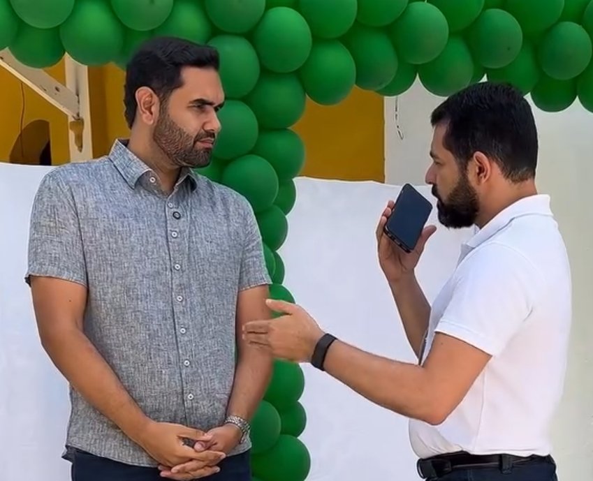 O prefeito de Piranhas, Tiago Freitas, de camisa cinza, concedendo entrevista de campo ao jornalista Ismael Alves, que segura um celular gravando o áudio sob um arco de balões verdes.