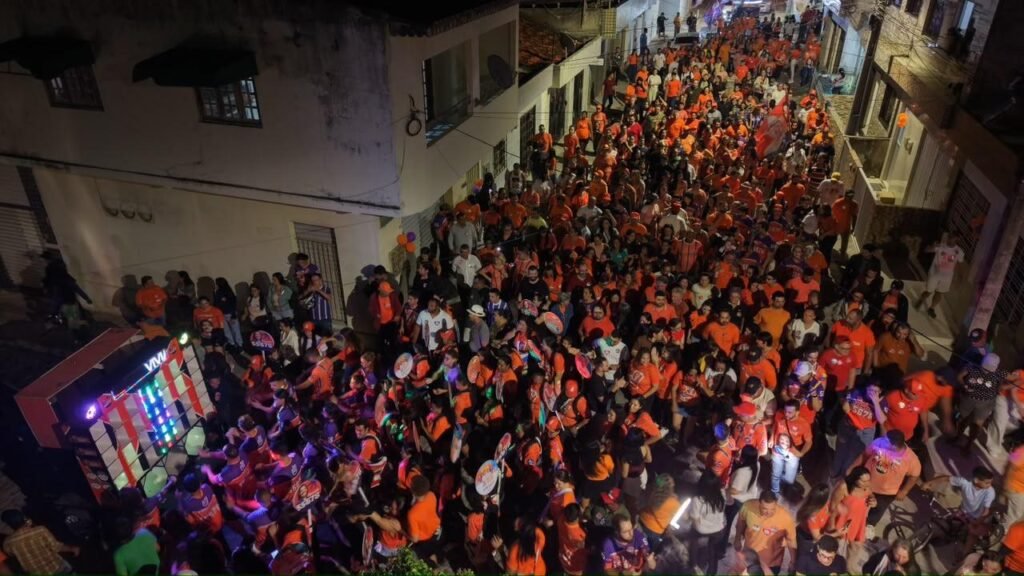 Vista aérea da multidão vestida de laranja lotando uma rua iluminada durante a caminhada de Viviane Facundes no bairro do Prado.