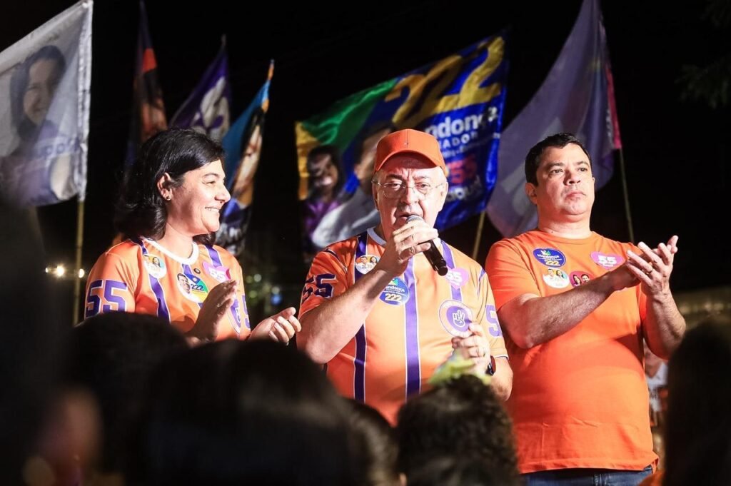 Viviane Facundes, o prefeito Joselito Gomes falando ao microfone e o vice-prefeito João Paulo de Lemos vestindo camisas laranjas durante ato político em Gravatá.