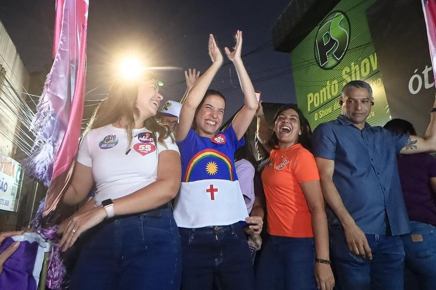 Viviane Facundes e Raquel Lyra comemoram sorrindo em caminhada política noturna em Caruaru. Raquel veste blusa com a bandeira de Pernambuco e braços erguidos. Viviane está à esquerda, de camisa branca com adesivos do número 55.