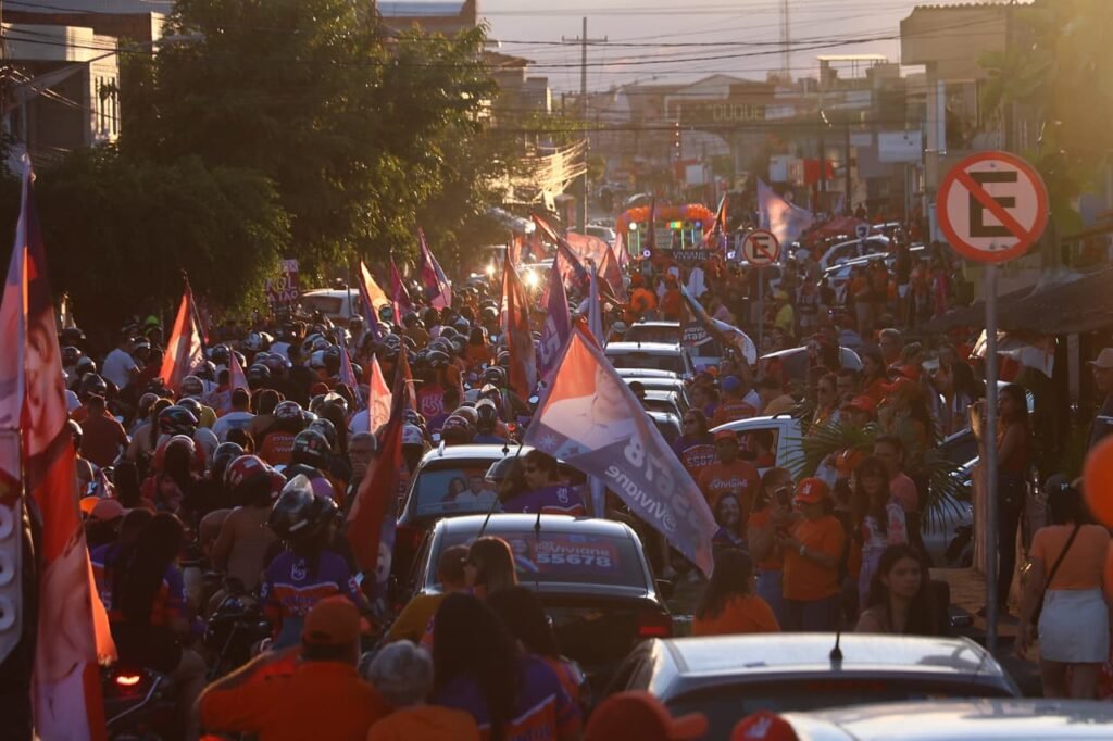 Fotografia externa em plano geral de uma movimentada carreata política pelas ruas de Gravatá, reunindo uma multidão de apoiadores, veículos, motos e bandeiras com as cores da campanha de Viviane Facundes para deputada estadual nas eleições de 2026.