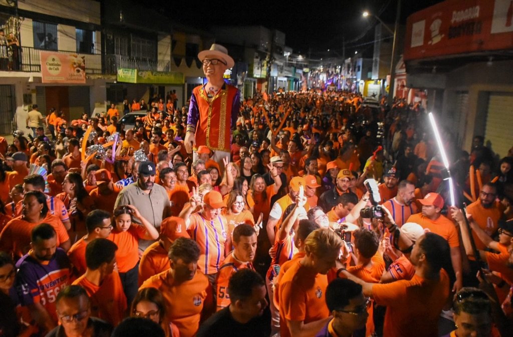 Multidão vestida de laranja acompanha a candidata Viviane Facundes em passeata noturna pelas ruas de Gravatá com boneco gigante em destaque.