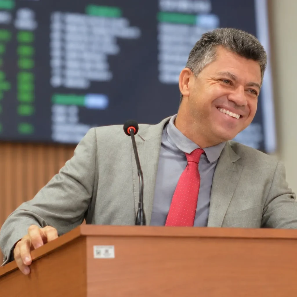 O vereador Ademir Batista sorri na tribuna da Câmara Municipal, vestindo terno cinza e gravata vermelha, com um painel eletrônico desfocado ao fundo.