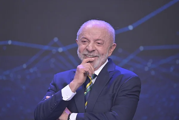 Fotografia de Luiz Inácio Lula da Silva sorrindo, vestindo terno escuro e gravata amarela, verde e azul, com a mão no queixo e fundo azul tecnológico com gráficos.