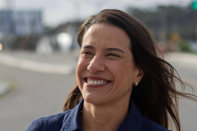 Retrato em close-up da governadora de Pernambuco, Raquel Lyra, sorrindo ao ar livre com o cabelo levemente ao vento e fundo urbano desfocado.
