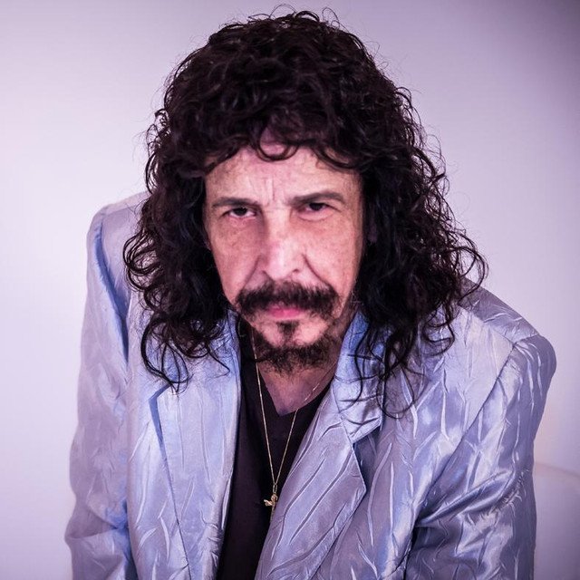 Retrato de homem com cabelos longos e cacheados, bigode, cavanhaque e paletó azul-claro sobre camisa preta.