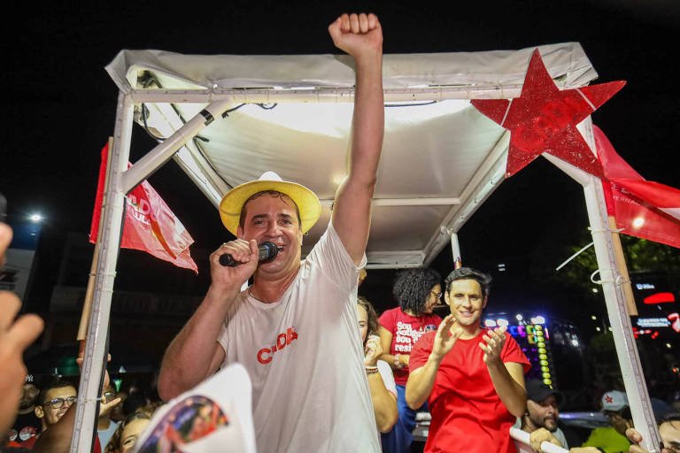 Fotografia de Cadu de Lula sorrindo em cima de um trio elétrico, vestindo camiseta branca e chapéu de palha amarelado, com o braço esquerdo erguido com o punho fechado e segurando um microfone na mão direita.