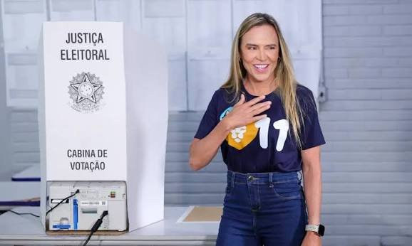 Fotografia de Celina Leão sorrindo ao lado de uma cabina de votação da Justiça Eleitoral, vestindo camiseta azul-marinho com o número 11 e calça jeans, com a mão direita no peito.