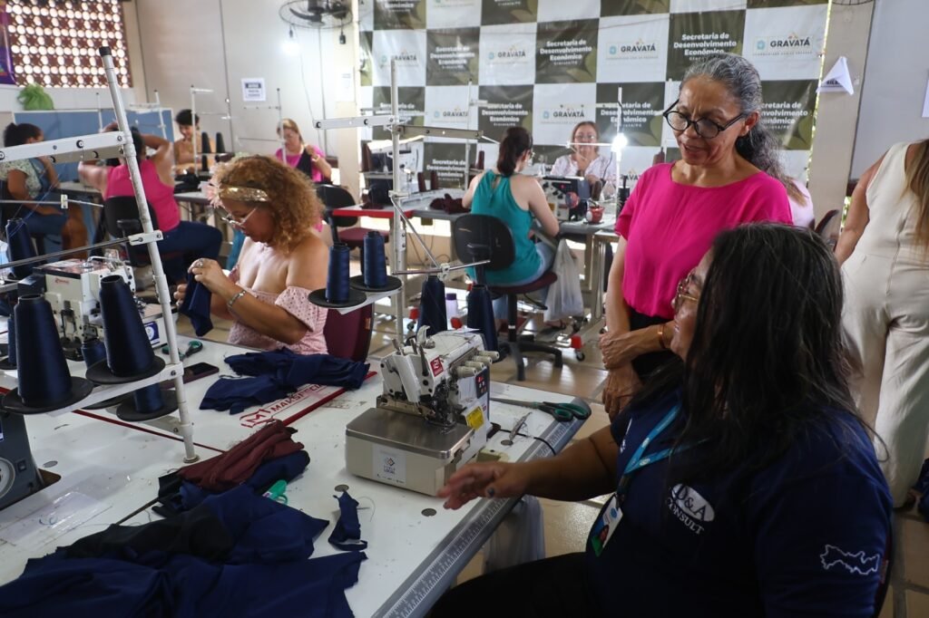 Fotografia interna colorida de um curso de costura em Gravatá, Pernambuco. No centro, duas mulheres conversam sobre técnicas de costura, com uma delas sentada diante de uma máquina de costura e a outra de pé ao lado. No fundo, várias outras mulheres são vistas trabalhando em máquinas de costura ao longo de fileiras de mesas, todas focadas em suas tarefas. Um banner ao fundo exibe logotipos do Governo de Gravatá e da Secretaria de Desenvolvimento Econômico.