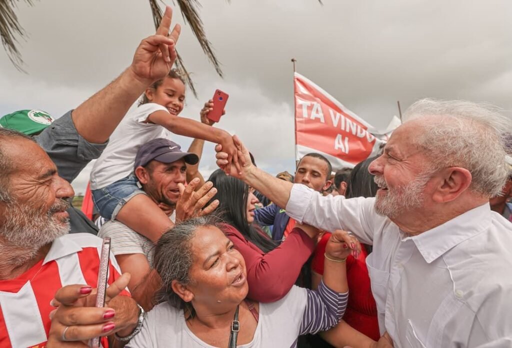 Fotografia de Luiz Inácio Lula da Silva cumprimentando apoiadores durante ato político. Lula sorri vestindo camisa branca enquanto segura a mão de uma criança nos ombros de um homem. Ao fundo, apoiadores seguram bandeira vermelha.