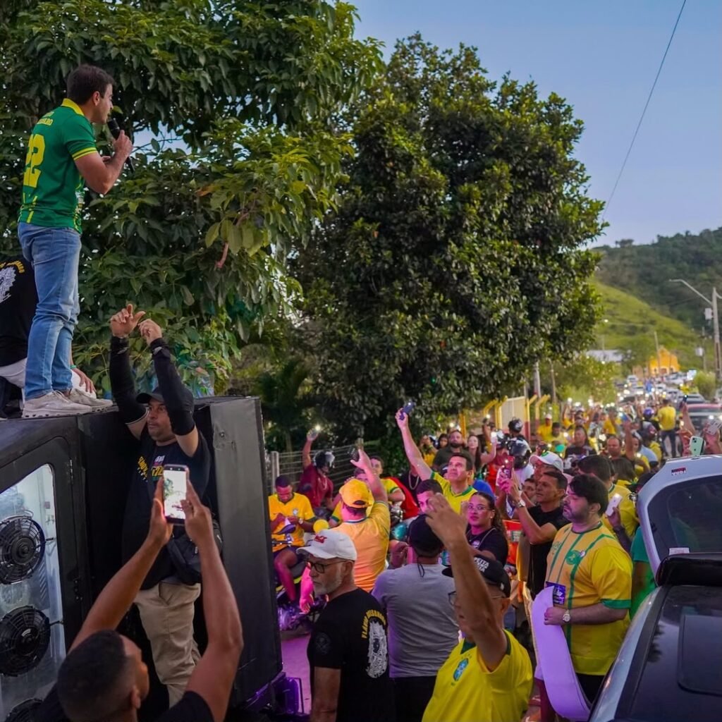 O deputado estadual eleito Gilson Machado Filho discursa em cima de um trio elétrico, vestindo camisa verde do Brasil com o número 22, durante carreata política pro-Flávio Bolsonaro em Chã Grande, Pernambuco. Uma multidão de apoiadores o assiste e registra com celulares