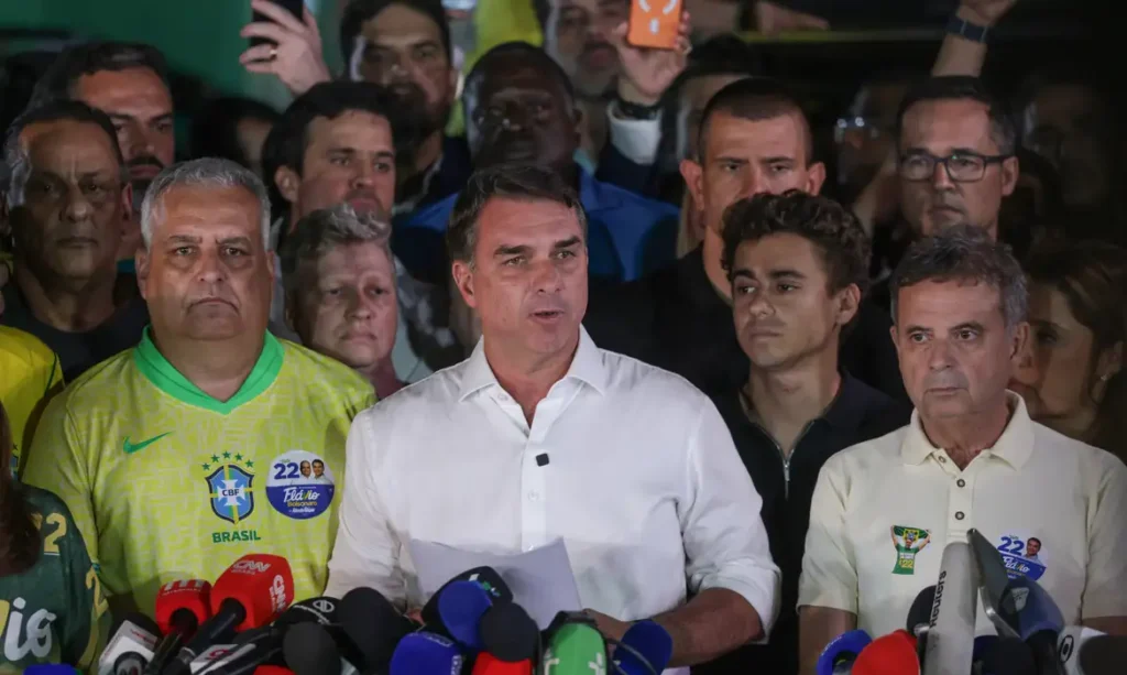Fotografia de Flávio Bolsonaro discursando em um pódio cercado por apoiadores e microfones de imprensa, vestindo camisa social branca e segurando folhas de papel.