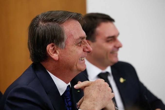 O ex-presidente Jair Bolsonaro sorri em primeiro plano com os dedos entrelaçados, enquanto o senador Flávio Bolsonaro aparece sorrindo ao fundo desfocado.