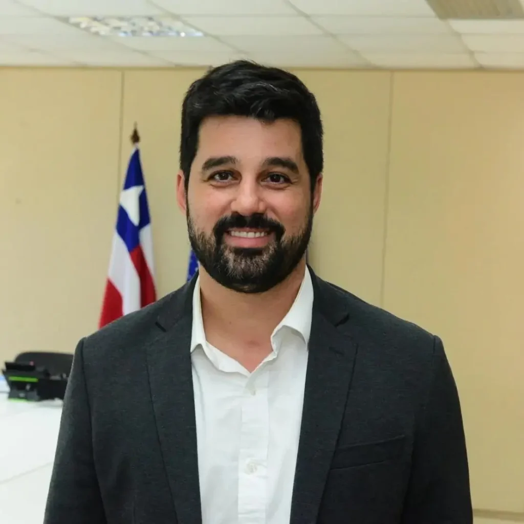 Foto oficial de Pablo Souza, secretário de Mobilidade de Salvador (Semob), usando blazer escuro e camisa branca, em frente à bandeira da Bahia