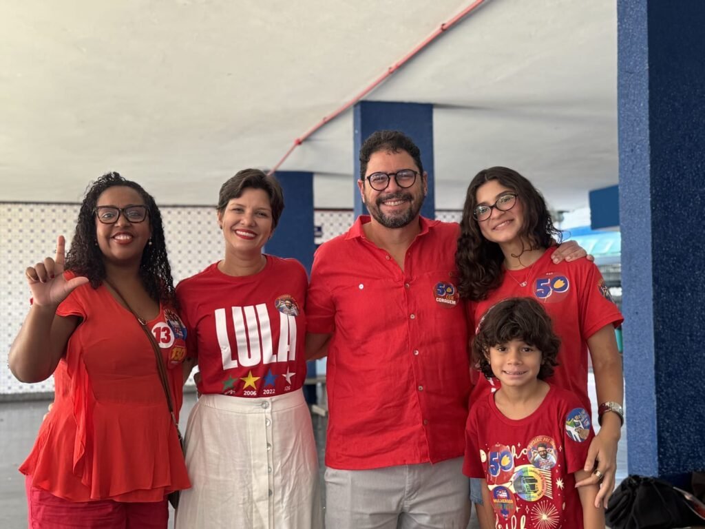O candidato Ivan Moraes, de camisa vermelha com o número 50, sorri ao lado da esposa, que usa camisa "LULA 2026", e dos filhos, em ambiente de votação com apoiadores fazendo o "L" com a mão