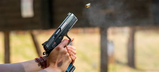 : Imagem em ângulo médio mostra as mãos de uma pessoa segurando uma pistola preta e prata no momento exato em que um tiro é disparado. Um estojo de bala deflagrado é visto saindo da arma, com um pouco de fumaça. O fundo está desfocado, sugerindo um estande de tiro ou ambiente externo.