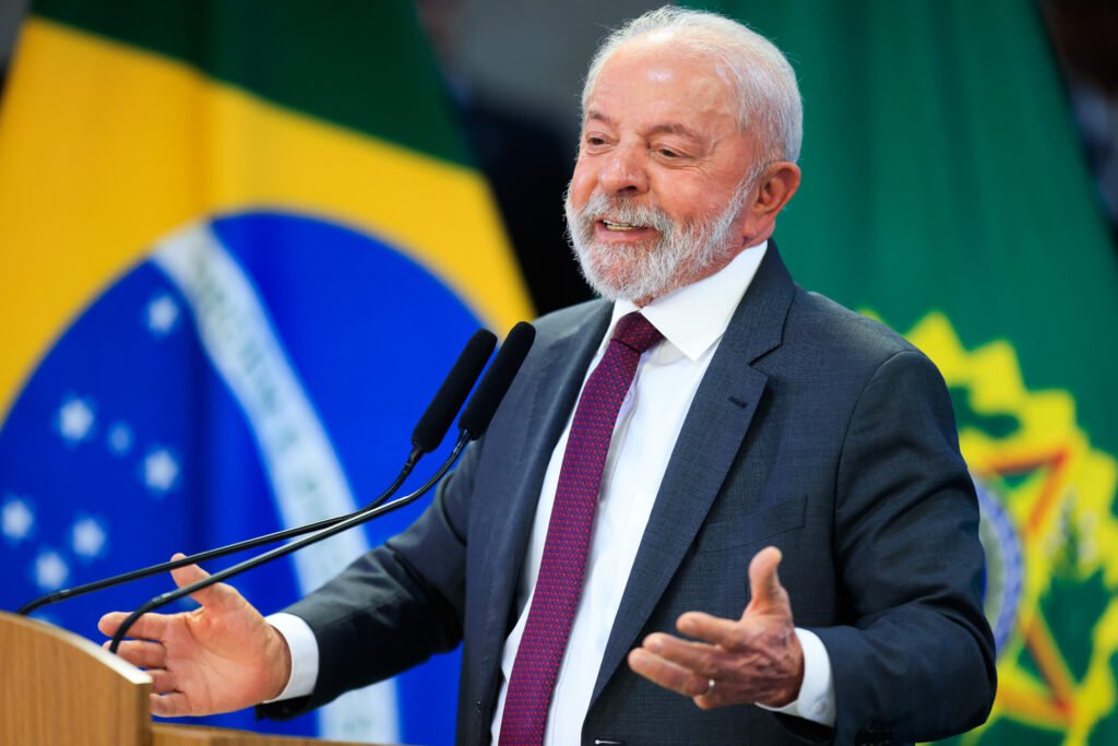 Fotografia em plano médio do presidente Luiz Inácio Lula da Silva discursando. Ele está atrás de um púlpito de madeira com microfones pretos, de terno cinza, camisa branca e gravata vermelha bordô estampada. Ele está de braços abertos e mãos estendidas, sorrindo e olhando para a direita. Ao fundo, as bandeiras do Brasil (à esquerda) e a bandeira presidencial verde (à direita).