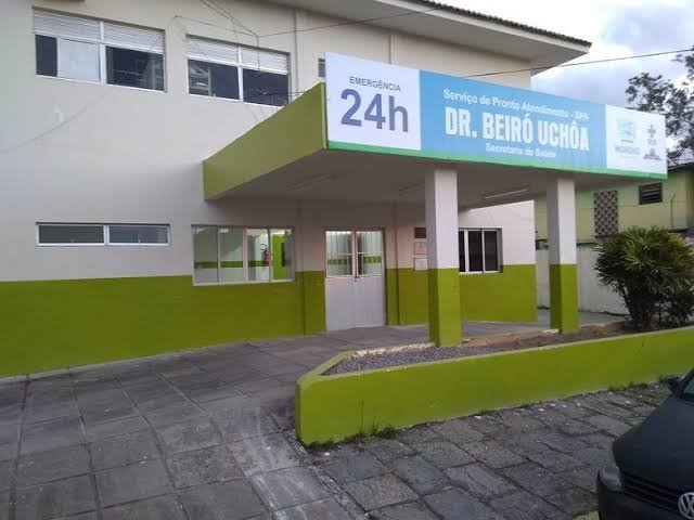 Fachada da Policlínica Doutor Beiró Uchôa, no município de Moreno, com destaque para a placa azul e branca do Serviço de Pronto Atendimento (SPA) Emergência 24h da Secretaria de Saúde.