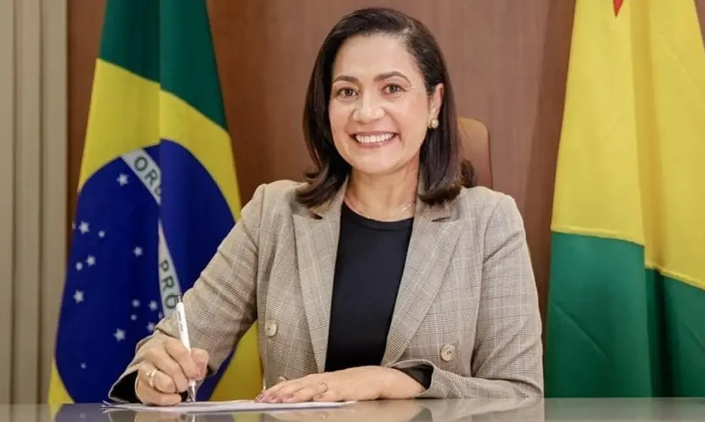 Fotografia oficial de Mailza Assis sorrindo, vestindo blazer xadrez bege sobre blusa preta, sentada à mesa assinando um documento entre as bandeiras do Brasil e do Acre.