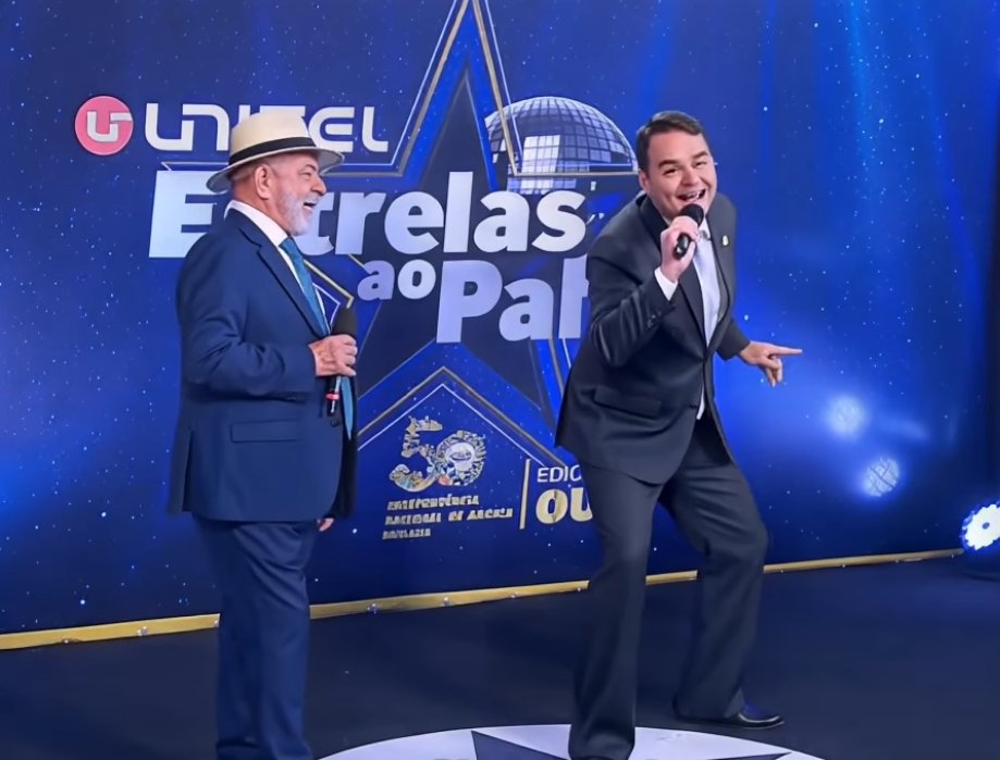 Meme gerado por inteligência artificial mostrando o presidente Lula e o senador Flávio Bolsonaro cantando juntos no palco do programa "Unitel Estrelas ao Palco".
