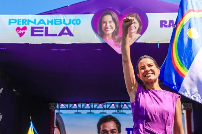 Raquel Lyra sorri e ergue o pugno fechado durante ato de campanha eleitoral, vestindo roupa roxa, ao lado da bandeira de Pernambuco e de faixa com a frase "Pernambuco com Ela