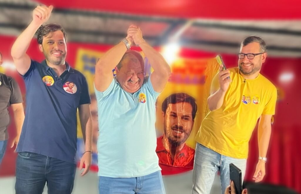 Nilson Pereira sorrindo com os braços levantados em celebração, ao lado de Gabriel Porto e Aglailson Victor, em evento político com banners ao fundo.