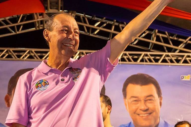 Fotografia de Omar Aziz com o braço esquerdo erguido em aceno, vestindo camisa polo rosa com adesivos eleitorais, sorrindo durante comemoração de campanha em palco.