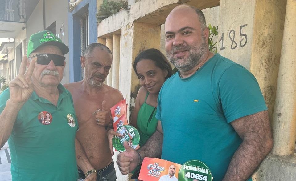 O candidato a deputado federal Pê Guerra posa sorridente ao lado de apoiadores e de um eleitor fazendo sinal com os dedos durante caminhada de campanha em Carpina.