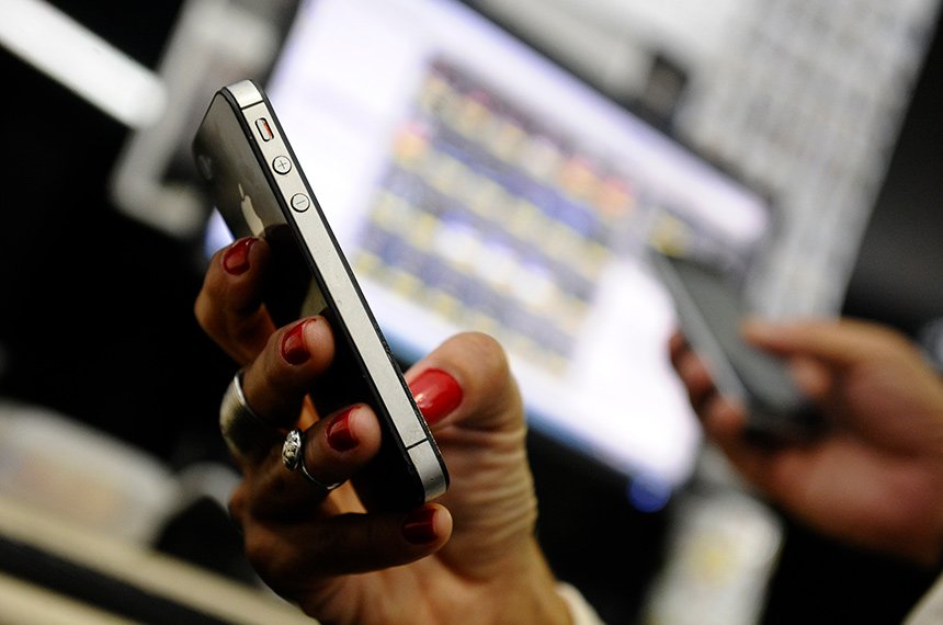 Mão com unhas pintadas de vermelho segurando um smartphone escuro, com um computador desfocado ao fundo, ilustrando a proibição de propaganda eleitoral e pedidos de voto nas redes sociais durante o dia das eleições.