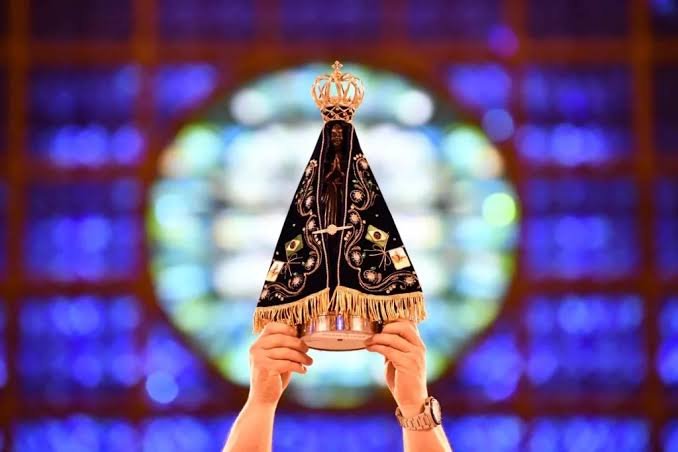 Imagem de Nossa Senhora Aparecida sustentada por mãos erguidas, com manto azul-escuro bordado e coroa dourada, diante de vitral iluminado.