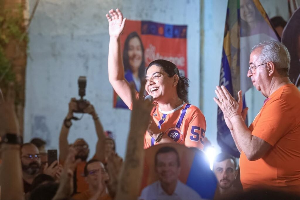 Viviane Facundes sorri e acena com o braço direito levantado, segurando um microfone, ao lado do prefeito Joselito Gomes durante evento de campanha política em Gravatá.