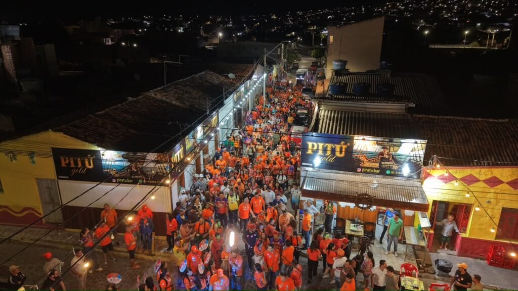 Vista aérea noturna de uma rua movimentada em Gravatá preenchida por apoiadores de Viviane Facundes vestidos com camisas laranjas durante caminhada porta a porta.