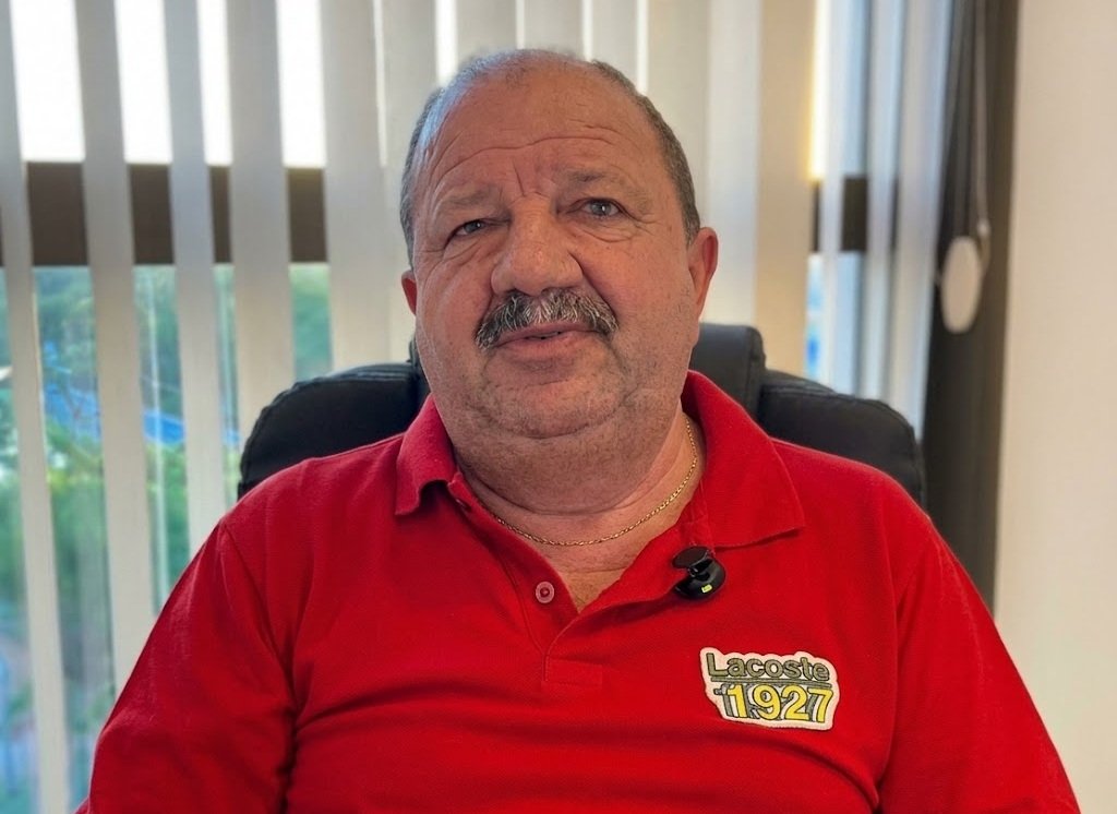 Retrato em plano médio de Nilson Pereira, primeiro suplente do PL para a Câmara do Recife, um homem de pele clara e bigode espesso, vestindo camisa polo vermelha com logomarca vintage "Lacoste 1927". Ele está sentado em uma cadeira de escritório em frente a uma janela com persianas verticais.
