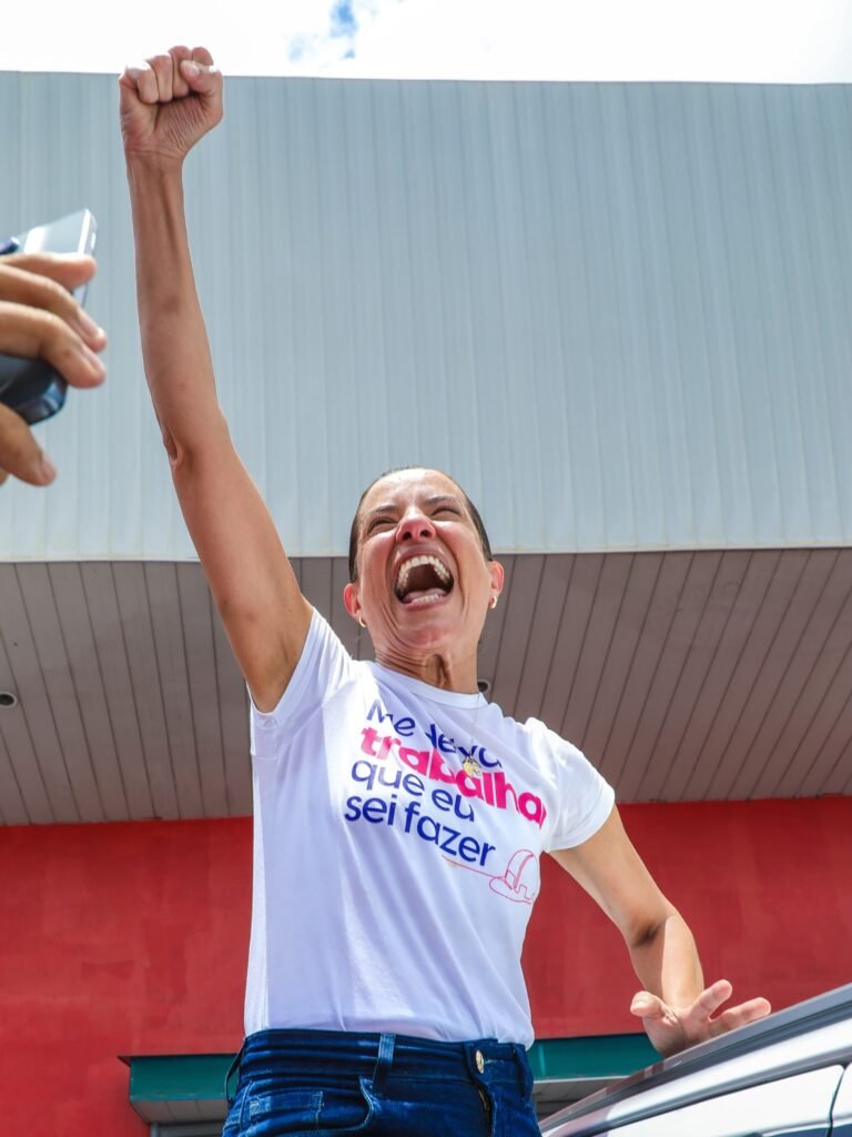 Raquel Lyra, reeleita governadora de Pernambuco no primeiro turno das eleições de 2026, comemora a vitória com o braço direito erguido e punho fechado, vestindo camiseta branca com a frase impressa "Me deixe trabalhar que eu sei fazer" e calça jeans.