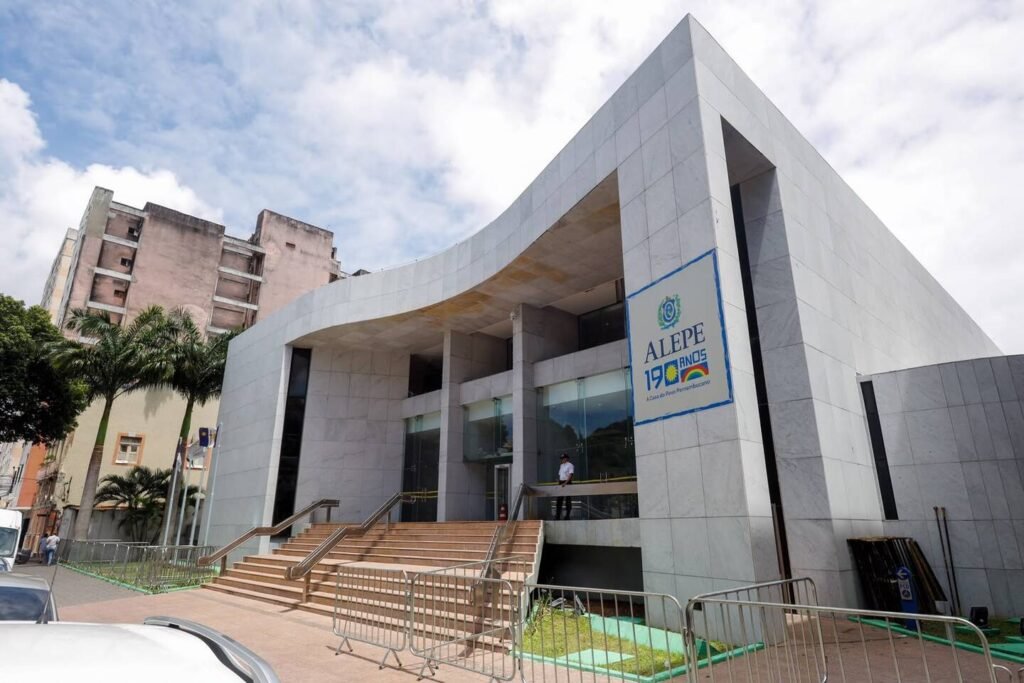 Fachada moderna da Assembleia Legislativa de Pernambuco (Alepe), revestida de blocos de mármore cinza claro, com escadaria frontal, portas de vidro e uma placa azul afixada na parede com a logomarca oficial da instituição.