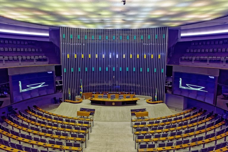 Plenário da Câmara dos Deputados em Brasília, com fileiras semicirculares de poltronas para parlamentares, a mesa diretora ao centro e grandes painéis eletrônicos nas laterais.