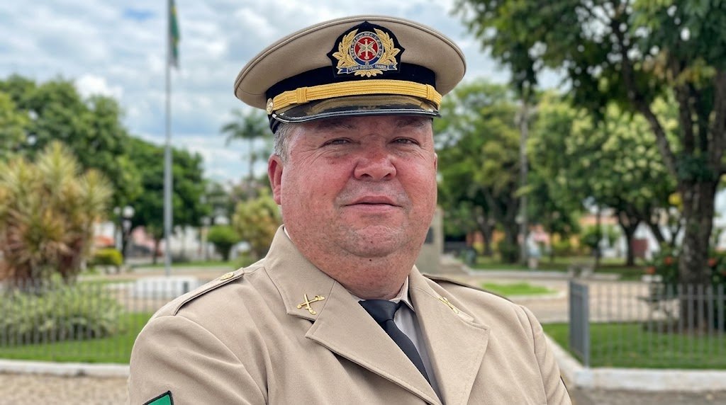 Retrato em primeiro plano do Sargento Venicius Rodrigues dos Santos, da Polícia Militar da Bahia (PMBA), fardado com uniforme bege de gala e quepe, sorrindo em uma praça arborizada em Vitória da Conquista.