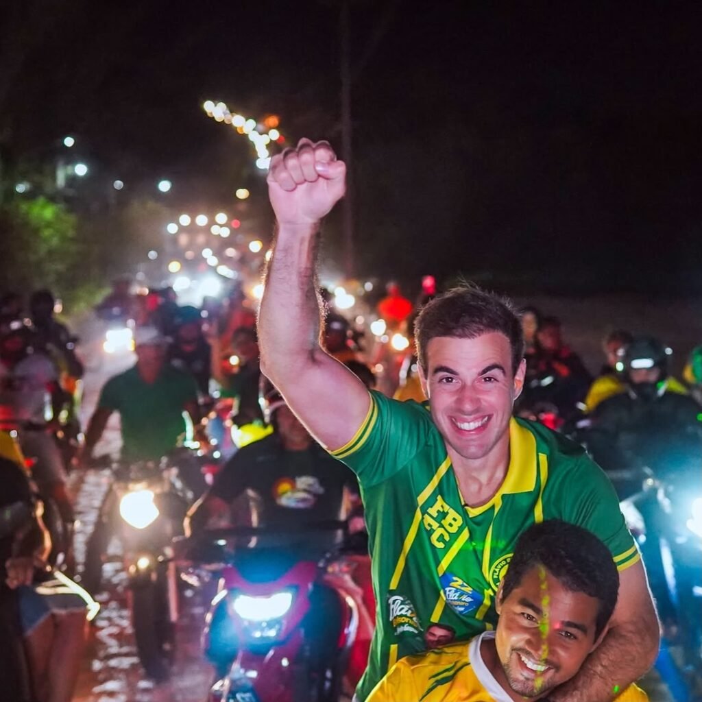 Alt Text: Close noturno sorridente de Gilson Machado Filho como garupa em uma moto, com o punho direito cerrado e erguido em sinal de vitória. Ele veste uma camisa verde e amarela com o número 22. O piloto também sorri e o fundo é uma carreata noturna com luzes desfocadas