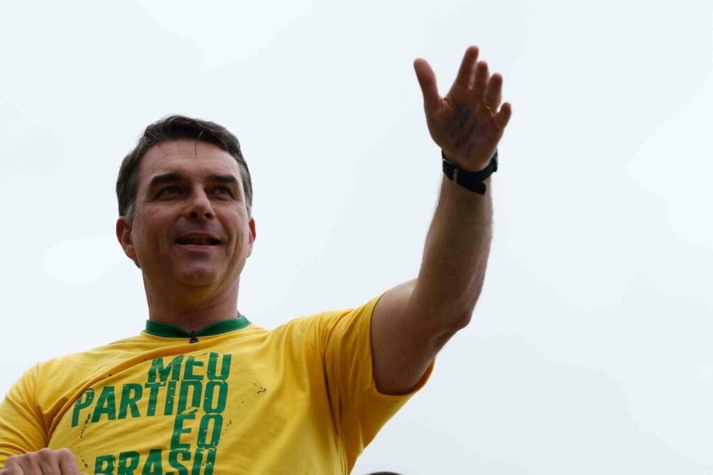 Alt Text: O senador e candidato à Presidência Flávio Bolsonaro sorri e acena com a mão esquerda erguida para eleitores em evento político. Ele veste uma camisa amarela com gola verde onde se lê a frase "MEU PARTIDO É O BRASIL".