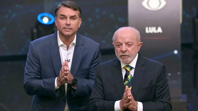 Fotomontagem em estúdio de debate eleitoral mostrando Flávio Bolsonaro (à esquerda, de fato escuro e camisa branca, com as mãos juntas em oração) ao lado de Luiz Inácio Lula da Silva (à direita, de fato escuro, gravata com as cores do Brasil e mãos juntas). Ao fundo, observa-se o logótipo da Band e a indicação de púlpito de debate.