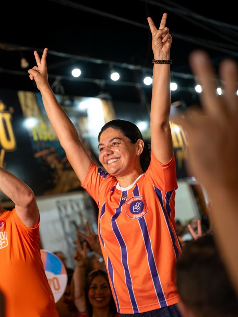 Foto de Viviane Facundes sorrindo, com os braços levantados e fazendo o sinal de 'paz e amor' com as duas mãos. Ela veste uma camisa de campanha laranja e listrada, com o logo 'Time Vivi 55678'. Ao fundo, uma multidão desfocada, luzes de festa e um cartaz parcialmente visível.